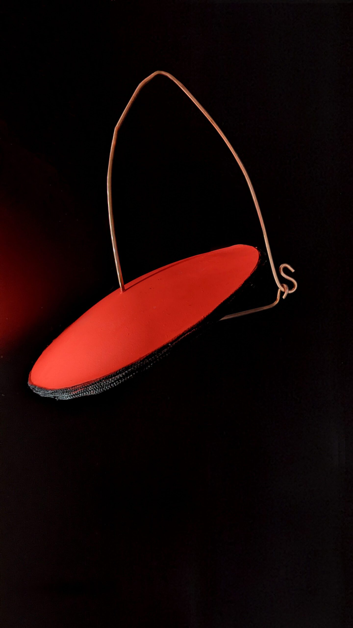 A UFO-like shape in neon orange/pink with a black corded edge, suspended from nothing by a steel wire frame on a black background.