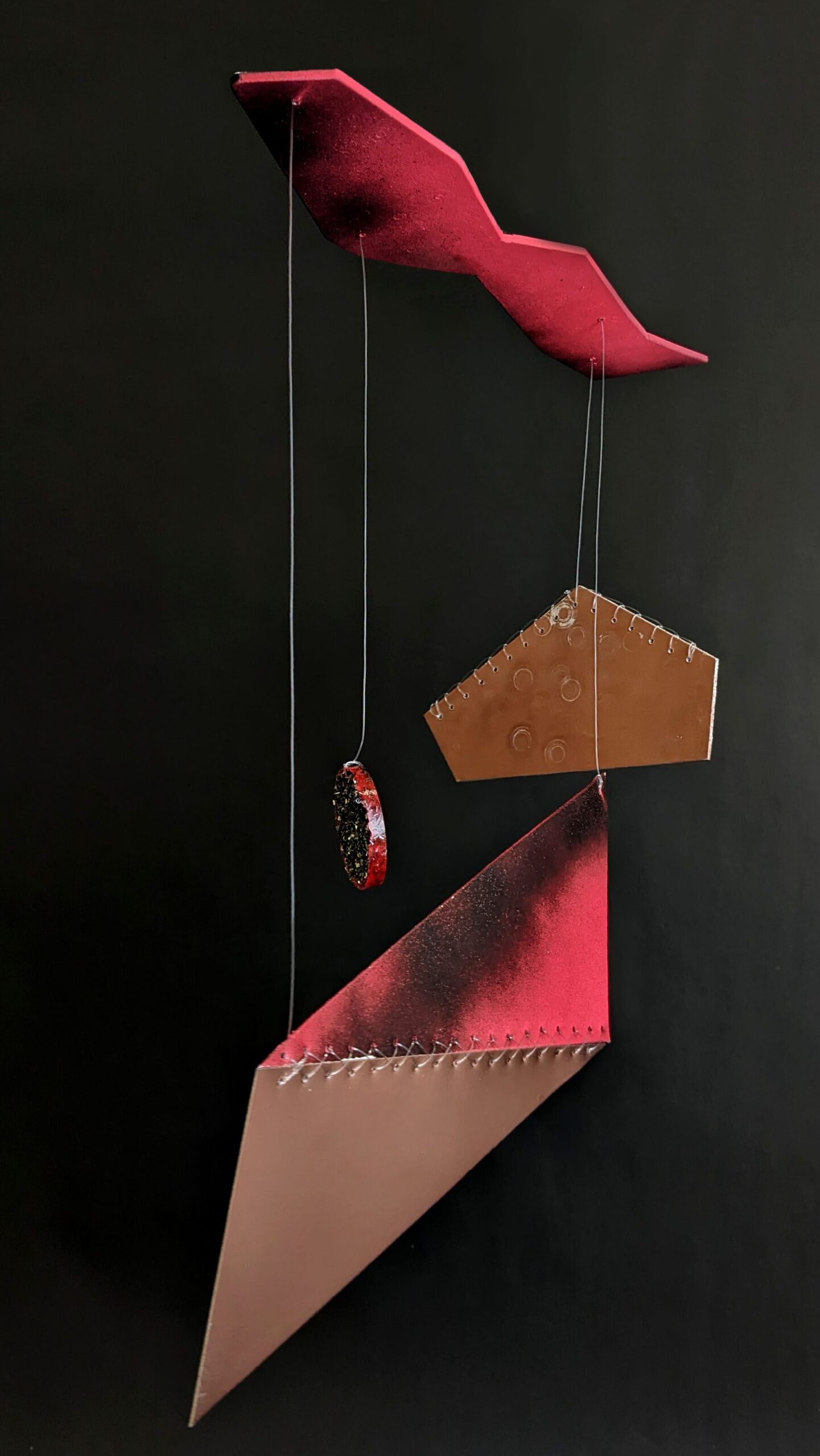A hanging sculpture  with three pendants, the largest a parallelogram made from two triangular aluminium sheets joined with what is the back of a traditional cross stitch, spray painted neon orange/pink, black and copper.  The second larges an irregular pentagon within monofilament blanket stitch across two of the edges with round scratched details. The final, and smallest, an oval pendant made from reclaimed steel shavings suspended in resin and paint. All are suspended from an irregularly shaped steel sheet also black and neon pink/orange. 